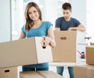 Young happy couple moving in their new house and unpacking boxes, she is carrying a carton box and smiling at camera